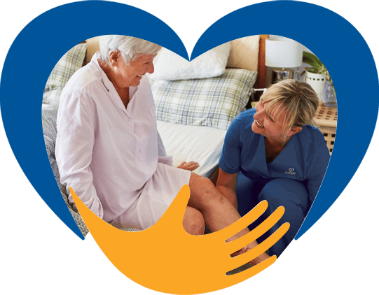 A woman helping an elderly woman sitting on a bed. Both are smiling.